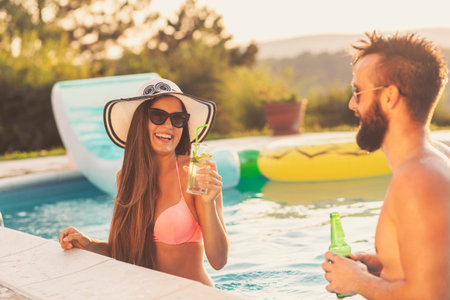 Couple in love at a poolside summer party, standing in water next to the swimming pool edge,...