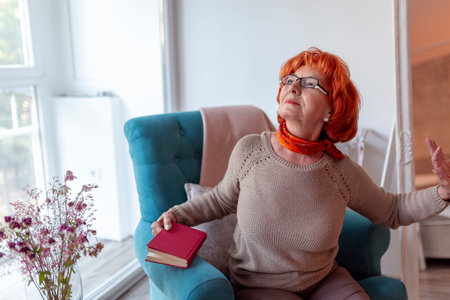Senior woman sitting in an armchair, stretching and yawning while reading a book