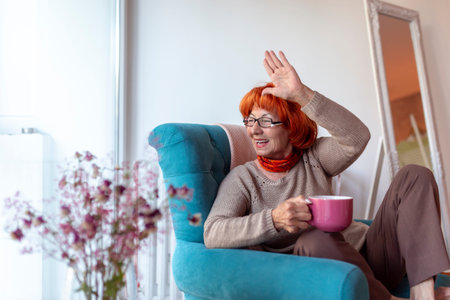 Beautiful senior woman sitting in an armchair by the window, drinking tea and waving to neighbors...