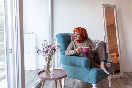 Beautiful senior woman sitting in an armchair by the window, drinking tea and looking out the window, relaxing at homeの写真素材