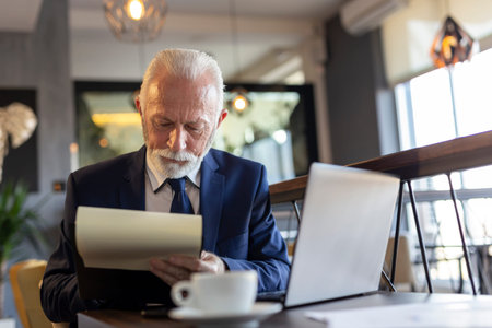 Senior businessman sitting at a restaurant table, working on a laptop computer, analyzing and...