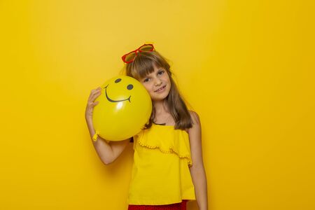 Portrait of a beautiful little girl holding and hugging a yellow balloon with a smiley face on it, isolated on yellow colored backgroundの写真素材