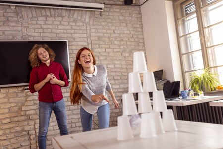Business people having fun at the office, playing office games while on a break, knocking down a pyramid of plastic cups by throwing a paper ballの写真素材