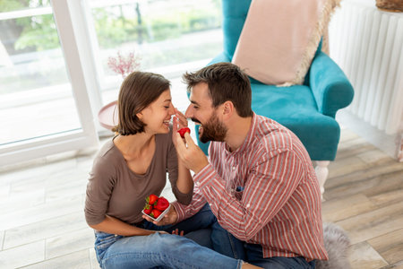 Couple in love sitting on the floor in their new home, eating strawberries, laughing and having fun
