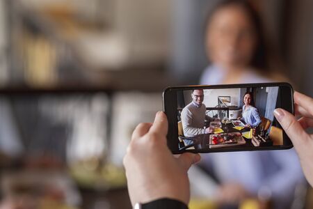 Couple in love having lunch at restaurant, celebrating their anniversary, being photographed by the waiter, making memories with smart phoneの写真素材