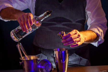 Bartender making a cocktail, pouring vodka from a bottle into a cocktail measuring cup, adding all...