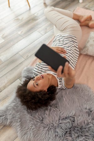 Top view of a beautiful young mixed race woman lying on the living room floor, taking a slefie with...