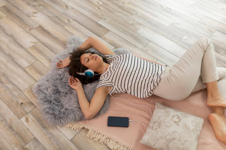 High angle view of beautiful mixed race woman lying on the floor, wearing a headset, listening to the music and relaxing at homeの写真素材