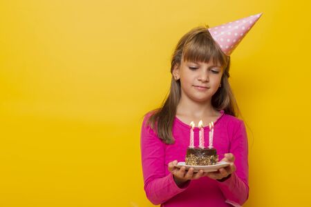 Beautiful little girl wearing birthday hat, holding a birthday cake with lit candles on it, making a wish and blowing candles, isolated on yellow colored backgroundの写真素材