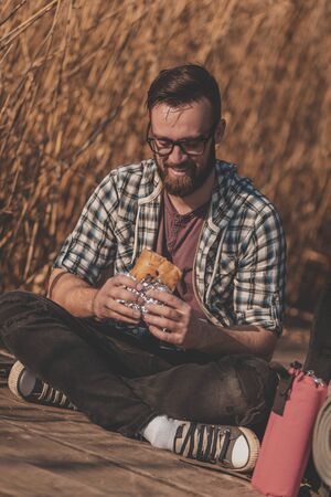 Male backpacker sitting on the wooden lake docks, taking a break from hiking, relaxing in nature and eating a sandwichの写真素材