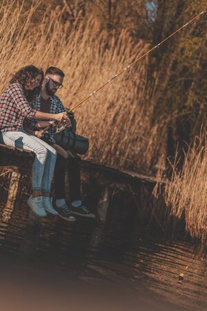 Couple in love sitting at lake docks, fishing and enjoying a beautiful sunny autumn day in natureの写真素材