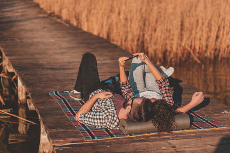 Beautiful couple in love lying on a picninc blanket at lake docks, taking a selfie and having fun on...