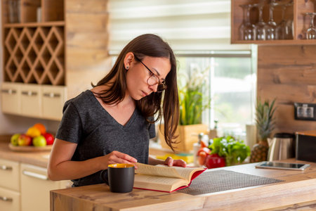 Beautiful young woman sitting at kitchen counter in the morning, drinking coffee and reading a book,...