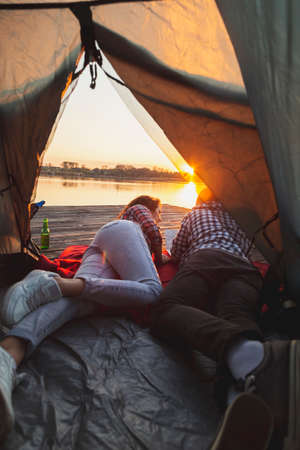 Young couple in love camping on lake docks, lying on the tent entrance, enjoying beautiful sunset and reading the mapの写真素材