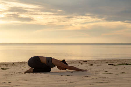 Young woman training on the beach early in the morning, practising yoga and stretching out, holding Balasana or child's poseの写真素材