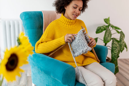 Beautiful young mixed race woman sitting in an armchair,  knitting a scarf and relaxing at home