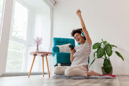 Beautiful young mixed race woman relaxing at home, sitting in an armchair and eating a fresh croissant for breakfastの写真素材