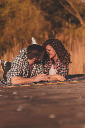 Beautiful couple in love lying on a picninc blanket at lake docks, taking a selfie and having fun on a sunny autumn dayの写真素材