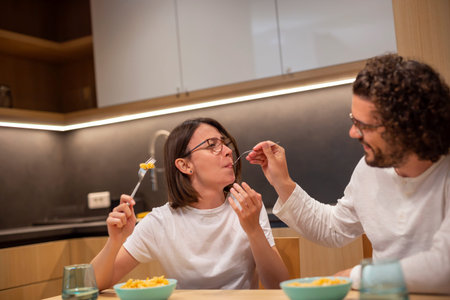 Couple in love wearing pajamas, sitting at kitchen table and having fun while eating dinner at home,...