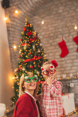 Beautiful mother and daughter sitting by nicely decorated Christmas tree, having fun at home on Christmas day, trying on Christmas party glassesの写真素材