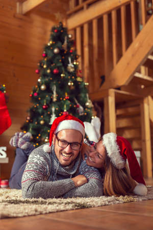 Top view of beautiful couple in love, wearing Santa hats, lying on the carpet next to a nicely decorated Christmas tree and enjoying Christmas morning at homeの写真素材