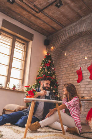 Beautiful young couple in love sitting by the nicely decorated Christmas tree, celebrating Christmas at home, drinking coffee and enjoying their time togetherの写真素材