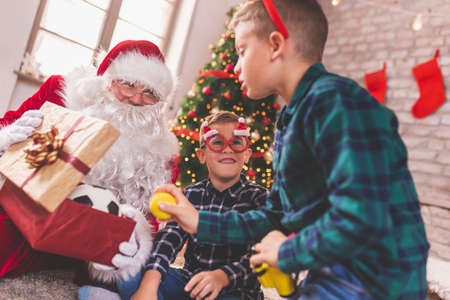 Two little boys sitting by nicely Christmas tree with Santa Claus, getting Christmas presents and having fun at home playing with their new toysの写真素材