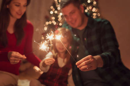 Mother, father and daughter celebrating Christmas Eve at home, sitting on the floor by nicely decorated Christmas tree, having fun while playing and waving with sparklersの写真素材