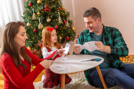 Beautiful young couple playing with their little daughter, holding big cardboard snowflakes and having fun while making Christmas decorations at homeの写真素材