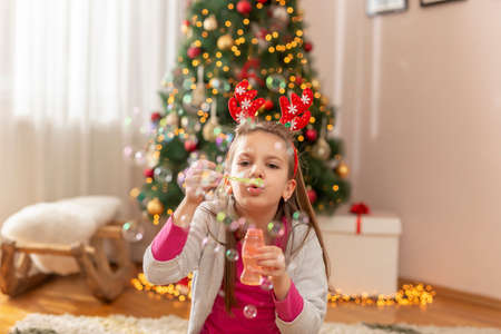Child having fun celebrating Christmas day at home, sitting near nicely decorated Christmas tree and blowing party whistleの写真素材