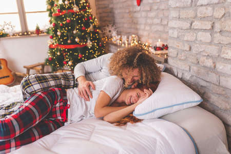 Young couple in love wearing pajamas lying in bed on Christmas morning, cuddling and relaxing at home during winter holiday seasonの写真素材