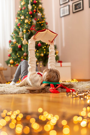 Beautiful little girl wearing costume reindeer antlers sitting next to nicely decorated Christmas tree, holding nicely wrapped Christmas presentの写真素材