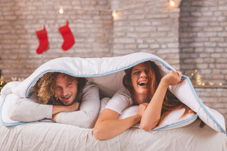 Young couple in love wearing pajamas lying in bed on Christmas morning, hugging, cuddling and relaxing at home during winter holiday seasonの写真素材