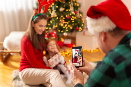 Man wearing Santa hat taking photos of his beautiful wife and daughter while celebrating Christmas...