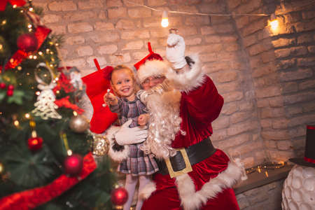 Beautiful little girl enjoying Christmas Eve while playing with her father wearing Santa Claus costume, holding candy cane while standing next to nicely decorated Christmas treeの写真素材