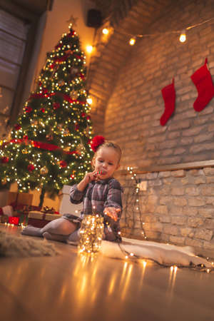 Beautiful little girl having fun at home on Christmas Eve, sitting on the carpet next to a nicely decorated Christmas tree, eating candy cane and playing with Christmas lightsの写真素材