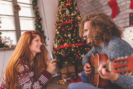 Beautiful young couple in love lying on the floor next to nicely decorated Christmas tree, having fun celebrating Christmas together at home, blowing party whistlesの写真素材