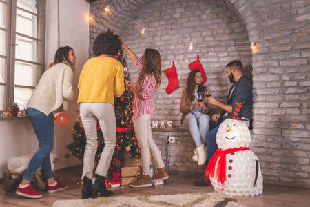 Group of young friends sitting by nicely decorated Christmas tree, celebrating Christmas at home, having fun while exchanging and opening presentsの写真素材