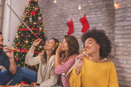 Group of young friends sitting by nicely decorated Christmas tree, celebrating Christmas at home, having fun while exchanging and opening presentsの写真素材