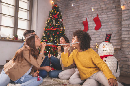 Group of young friends sitting by nicely decorated Christmas tree, celebrating Christmas at home,...