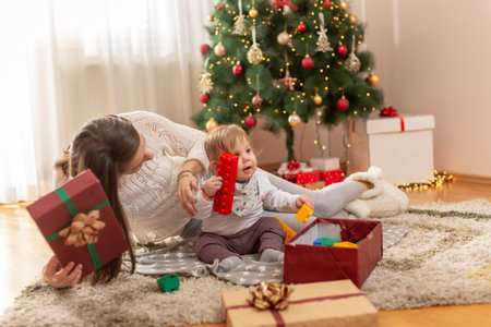 Mother and son having fun at home on Christmas morning, sitting by nicely decorated Christmas tree,...
