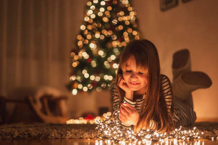 Beautiful little girl wearing costume reindeer antlers, sitting near nicely decorated Christmas tree, holding sparklers and having fun while celebrating New Year's Eve at homeの写真素材