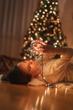 Beautiful young woman lying on the floor by nicely decorated Christmas tree, relaxing at home and enjoying winter holoday seasonの写真素材
