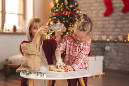 Mother and daughter sitting by nicely decorated Christmas tree, kneading dough for gingerbread Christmas cookies and having funの写真素材
