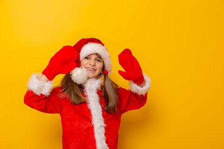 Beautiful little girl dressed as Santa Claus waving towards the camera and smiling, isolated on yellow colored background with copy spaceの写真素材