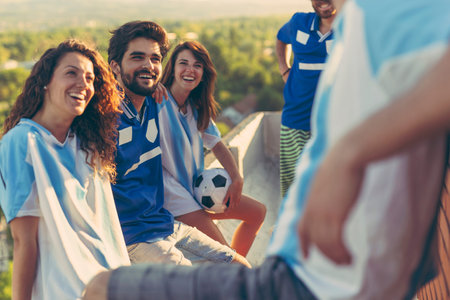 Group of football fans standing on a building rooftop terrace, hanging out and speaking, before...