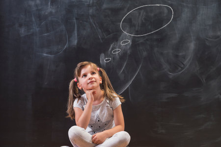 Beautiful little elementary school girl sitting in front of a chalkboard pensive and serious, thinking, with clouds drawn above her headの写真素材