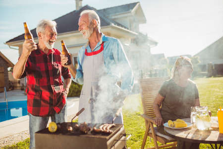 Senior couple having a backyard barbecue party with friends, grilling meat and having fun spending leisure time outdoorsの写真素材