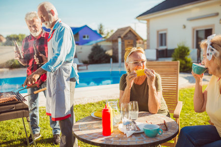Group of senior friends having a backyard barbecue party by the swimming pool, gathered around the...