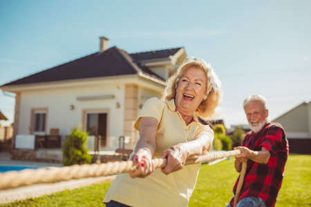 Senior couple having fun playing tug of war game, spending sunny summer day outdoors; group of elderly friends participating in rope pulling competition at retirement homeの写真素材